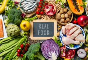 Variedad de comida real: vegetales, frutas, pescado y pollo alrededor de un letrero que dice "Real Food"
